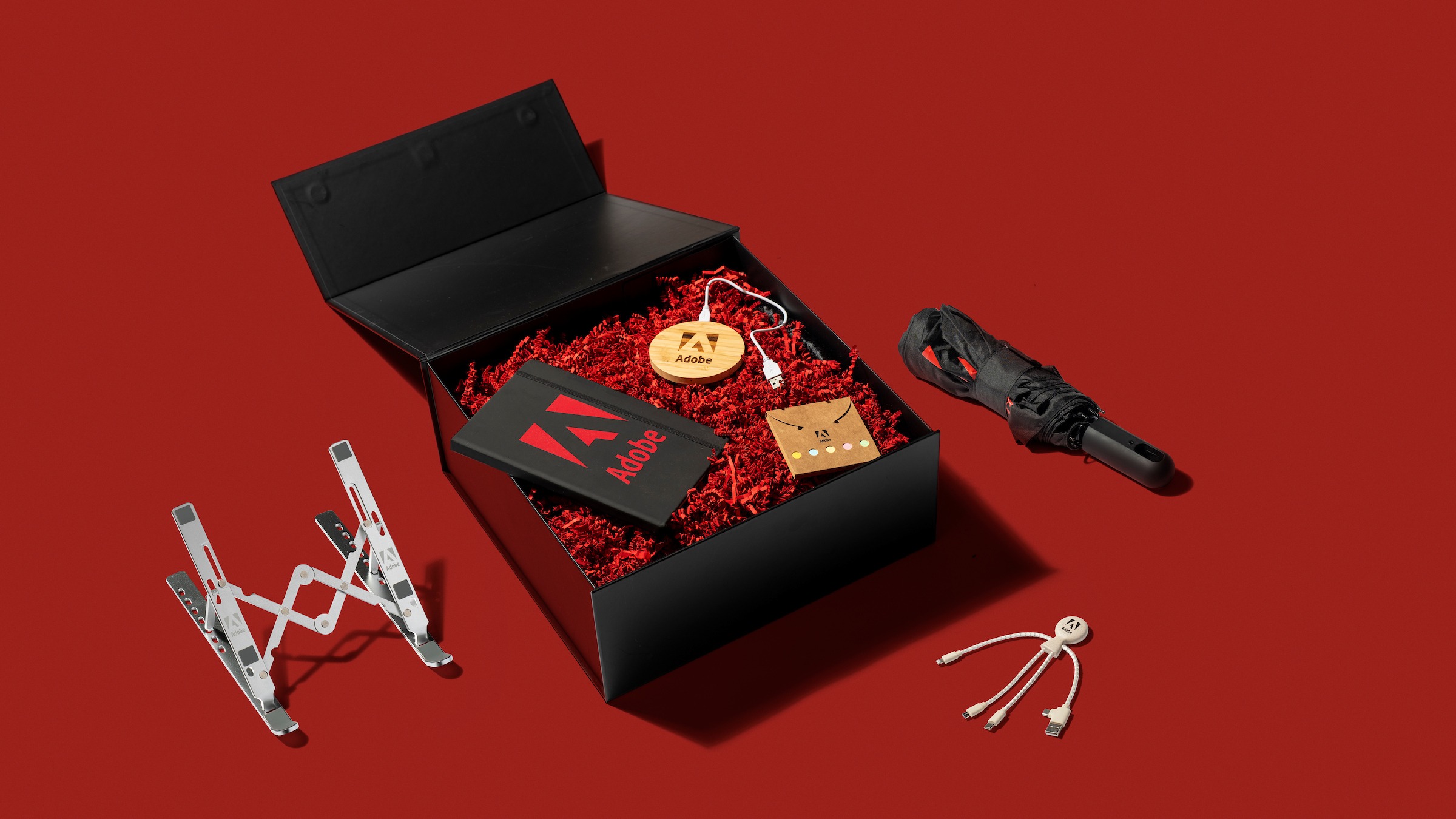 Flat lay of the Adobe Employee box; a black box with a laptop stand, notebook, phone charger pad, multi-usb stick and an umbrella, all on a dark red background